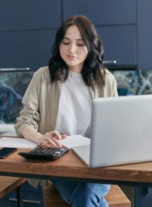 A young woman calculating finances at home using a laptop and calculator, reflecting a focused budgeting process.