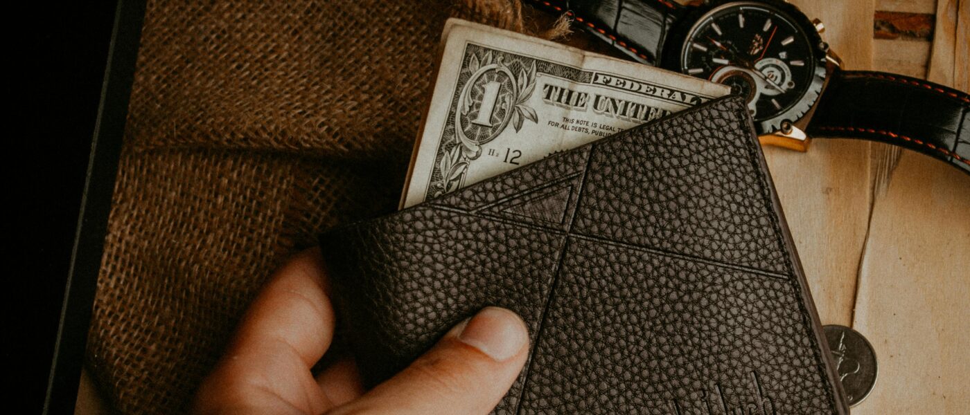 A hand holds a leather wallet with a dollar bill exposed, next to a wristwatch on a wooden surface.