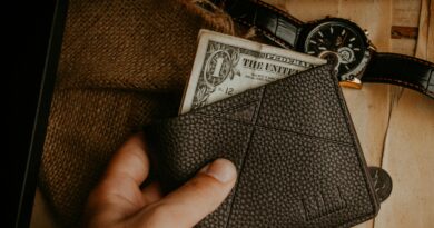 A hand holds a leather wallet with a dollar bill exposed, next to a wristwatch on a wooden surface.