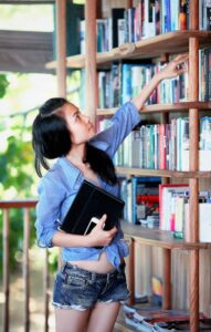 A young woman reaches for a book on a library shelf, holding a tablet.