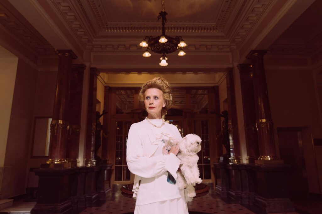 An elegant woman in a classic white dress holding a Maltese dog in a luxurious, vintage lobby.