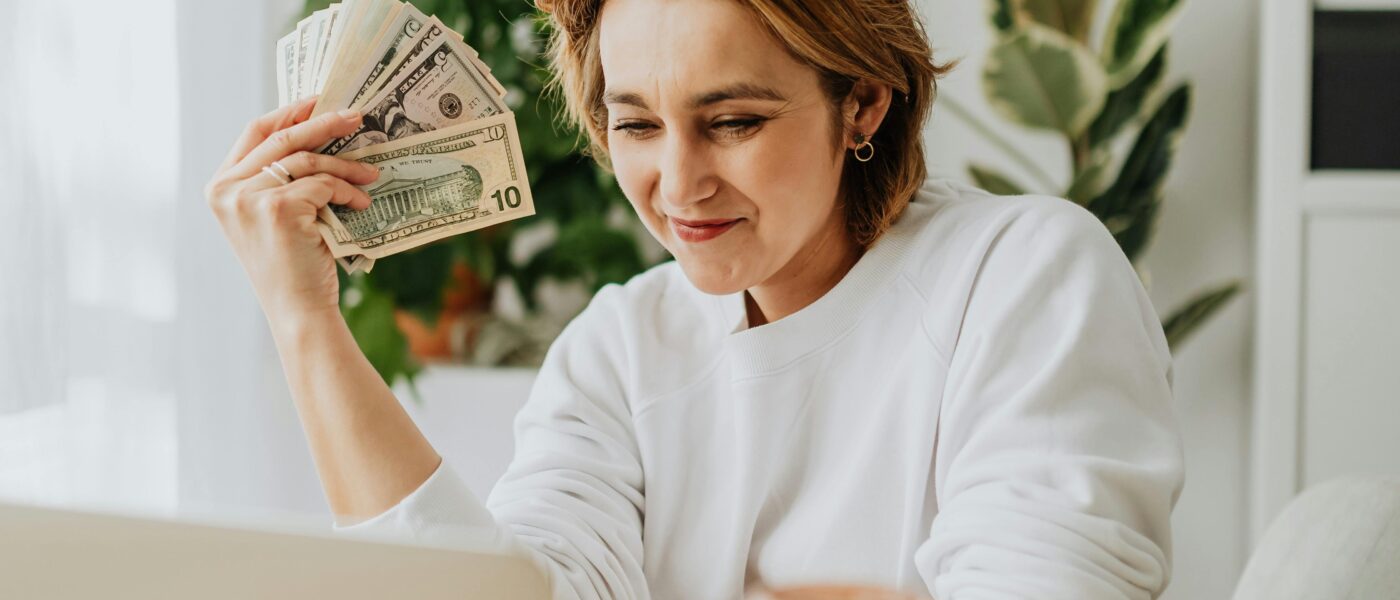 Woman seated at a desk holding dollar bills, working on a laptop, showcasing financial success from home.