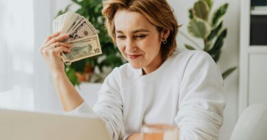 Woman seated at a desk holding dollar bills, working on a laptop, showcasing financial success from home.