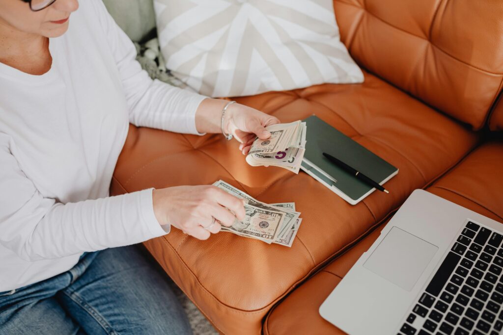 An adult woman sitting on a sofa, counting money with a laptop and notebook nearby, illustrating budget planning.