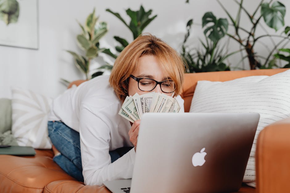 Woman holding cash while using a laptop on a cozy sofa at home, conveying financial success.