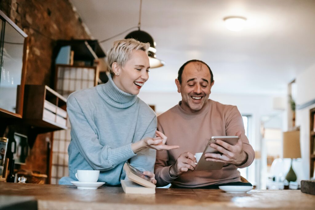 Cheerful adult diverse couple in casual clothes watching funny video on tablet and laughing while drinking coffee at table in apartment