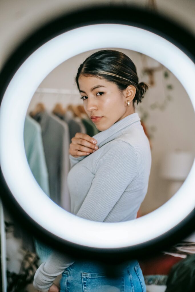 Confident young ethnic female in blouse and jeans standing in room near ring light and wardrobe and demonstrating trendy outfit