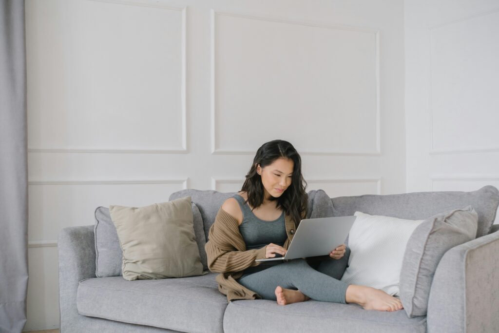 Young woman in active wear working from home on a gray couch with a laptop.