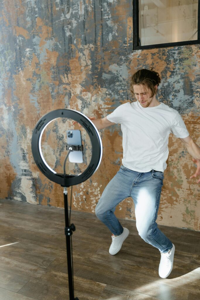 A young man dancing energetically while filming content indoors using a ring light.