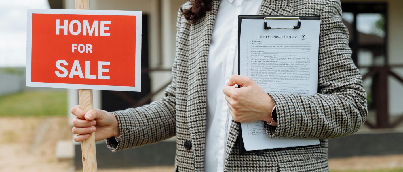 A real estate agent holding a home for sale sign and clipboard outside a property.