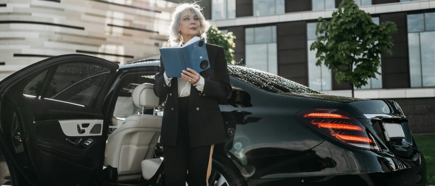 Senior businesswoman exits a black luxury car, holding documents near modern office building.